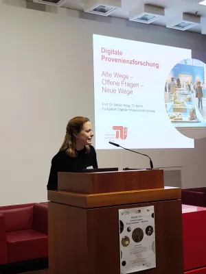 Meike Hopp steht hinter einem Podest auf der Bühne und spricht in ein Mikrofon. Hinter ihr befindet sich an der Wand eine Projektion der Vortragsfolien.